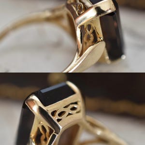 May include: Close-up of a gold ring featuring a large, rectangular black gemstone. The ring has intricate filigree detailing on the setting, and the band is a polished gold color. The ring is set against a blurred background.