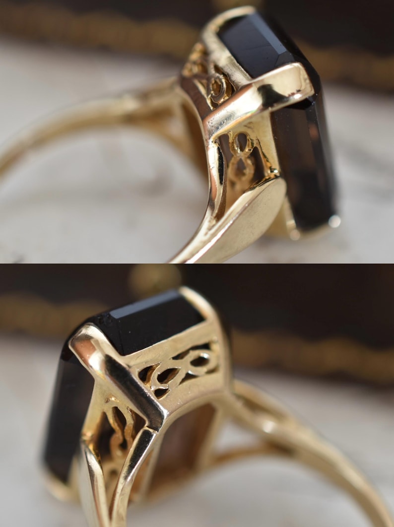 May include: Close-up of a gold ring featuring a large, rectangular black gemstone. The ring has intricate filigree detailing on the setting, and the band is a polished gold color. The ring is set against a blurred background.