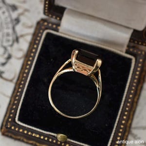May include: A gold ring with a large, rectangular, dark gemstone set in a detailed setting. The ring is displayed in a vintage jewelry box with a black velvet interior. The ring's band has a delicate, open design.