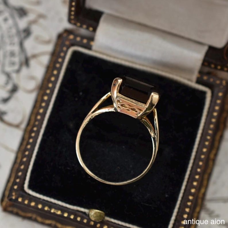 May include: A gold ring with a large, rectangular, dark gemstone set in a detailed setting. The ring is displayed in a vintage jewelry box with a black velvet interior. The ring's band has a delicate, open design.