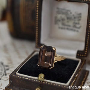 May include: A gold ring with a large, rectangular, smoky quartz gemstone. The ring is set in a vintage-style setting and is displayed in a small, antique jewelry box with a black velvet interior. The box is brown with gold accents.