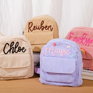 Könnte beinhalten: Vier Cord-Rucksäcke in Beige, Hellbraun, Lavendel und Rosa, jeweils mit einem Namen in einer Kontrastfarbe bestickt. Der lavendelfarbene Rucksack hat Blumenakzente und einen Regenbogen. Die Rucksäcke haben eine Vordertasche und verstellbare Träger.