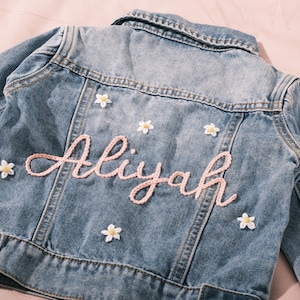 May include: Light blue denim jacket with the name "Aliyah" embroidered in pink script. The jacket is decorated with small white and yellow flower appliqués. A classic, personalized piece.