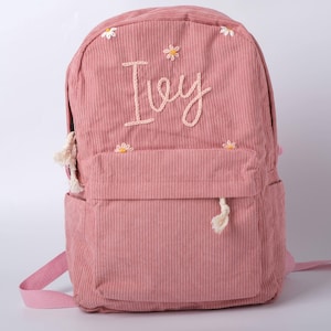 Op de afbeelding: Roze corduroy rugzak met de naam "Ivy" geborduurd. De rugzak heeft kleine madeliefjesaccenten en een voorvak. Verstelbare bandjes en een handvat maken het ontwerp compleet.