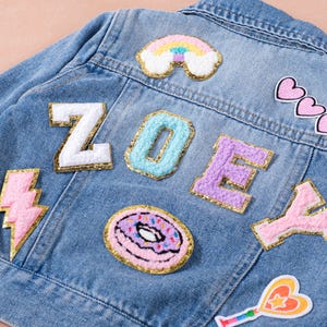 May include: A light blue denim jacket adorned with colorful patches. The patches include the name "ZOEY" in individual letter patches, a rainbow, hearts, a lightning bolt, a donut, and a star-shaped wand.