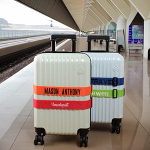 May include: Two upright rolling suitcases with vertical ridges and black handles. The front suitcase is cream with an orange strap reading MASON ANTHONY and a red strap reading Hasselquist. The back suitcase is white with blue and green straps.