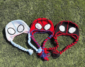 May include: Three crocheted beanies with earflaps and braided tassels. The first is white with light blue accents and white eye patches. The second is red with black webbing and white eye patches. The third is black with red accents and white eye patches.