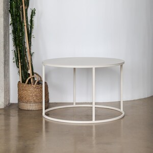 Op de afbeelding: Een ronde, crèmekleurige bijzettafel met een metalen frame. De tafel heeft een cirkelvormige basis en een bijpassend tafelblad. Links van de tafel staat een hoge groene plant in een gevlochten mand.