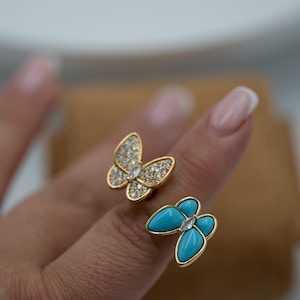 Op de afbeelding: Twee vlinderringen. Eén ring heeft een goudkleurige vlinder met heldere kristallen. De andere ring heeft een goudkleurige vlinder met turkooizen stenen en een helder kristal in het midden.