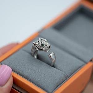 Op de afbeelding: Zilverkleurige ring met een jaguarhoofdontwerp, bezet met kleine, heldere stenen en groene ogen. De open ring ligt in een grijs fluwelen, oranje doosje.