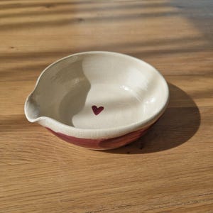 Könnte beinhalten: Eine handgefertigte Keramikschale mit Herzmuster. Die Schale hat einen cremefarbenen Innenraum und einen weinroten Rand. Ein kleines weinrotes Herz ist in der Mitte aufgemalt. Die Schale hat einen Ausguss auf einer Seite.