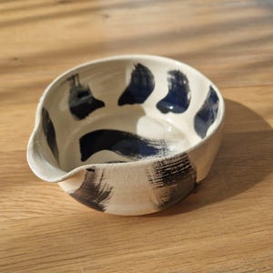 Könnte beinhalten: Eine handgefertigte Keramikschale mit Ausguss. Die cremefarbene Schale hat dunkelblaue Pinselstriche und Akzente. Das Innere hat dunkelblaue abstrakte Formen. Die Schale steht auf einer Holzoberfläche.