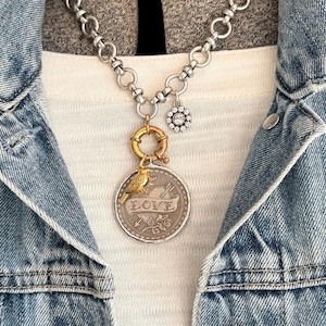 May include: Silver-tone chain necklace with a pendant featuring a gold bird perched above the word "LOVE" on a coin-shaped charm. Additional charms include a gold ring and a small, sparkling flower. The necklace is worn over a white shirt and a denim jacket.