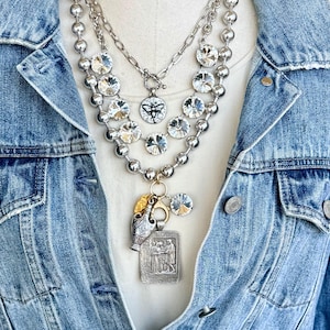 May include: A layered necklace featuring silver-toned ball chains, clear faceted crystals, and a toggle clasp with a circular bee charm. A central pendant cluster includes a small gold-toned leaf, a silver-toned amphora, and a rectangular metal plaque.