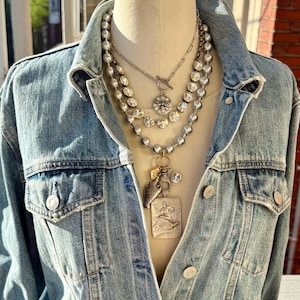May include: A light blue denim jacket worn over a mannequin, showcasing a layered necklace set. The necklaces feature various silver chains, sparkling crystals, and unique pendants. The jacket has buttoned pockets and a classic collar.