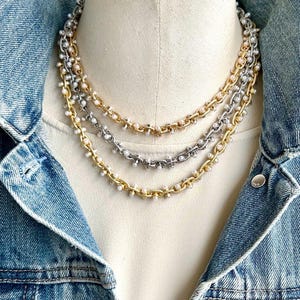 May include: Three layered necklaces with gold and silver chains, adorned with small white pearls. The necklaces are displayed on a mannequin, set against a denim jacket background. The chains have a delicate, feminine design.
