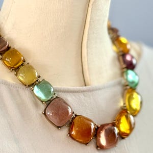 May include: A necklace featuring a series of square, translucent, multi-colored cabochon stones in shades of amber, yellow, green, and mauve, set in a metallic frame that rests against a cream-colored fabric surface.