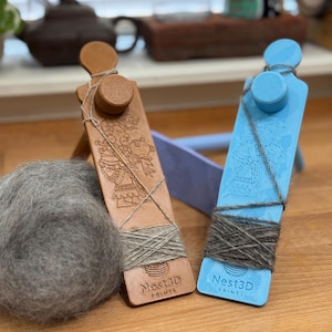 May include: Two yarn swift tools, one tan and one blue, with wound yarn. The tan tool has an etched design and the blue tool has a smooth surface. Both tools have the text "Nest3D Prints". A ball of grey yarn is in the foreground.