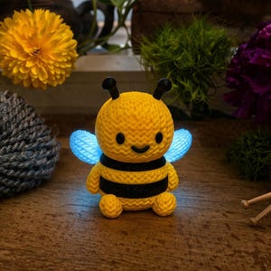 May include: A yellow and black crocheted bee with glowing blue wings. The bee has a smiling face and is surrounded by yarn and flowers. The bee is a handmade item.