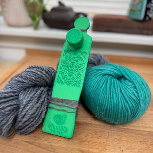 May include: A green yarn swift with a bee design and the text "Nest3D PRINTS" is displayed with two skeins of yarn. One skein is teal, and the other is grey. The yarn swift is wrapped with grey yarn.