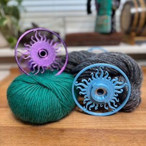 Puede incluir: Dos madejas de hilo, una verde y otra gris, sobre una superficie de madera, con dos herramientas de bobinado de hilo en forma de sol. Una herramienta es morada, la otra azul. Una cafetera y un barril de madera en el fondo.