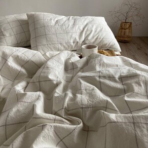 May include: White bedding set with a grid pattern. The set includes two pillows and a duvet cover, all with a subtle gray grid design. A white mug with a dark liquid sits on the bed, alongside a small snack.