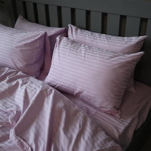 May include: A set of light purple bedding featuring a subtle, repeating vertical stripe pattern. The set includes two rectangular pillows and a matching duvet cover, all crafted from a smooth fabric with a soft, uniform lavender colour tone.