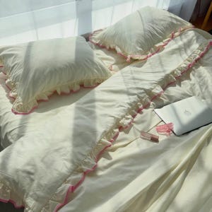 May include: A cream-colored bedding set featuring two pillows and a duvet cover, each adorned with ruffled edges trimmed in bright pink. The bedding is laid out flat, accompanied by a silver laptop, a small pink hair clip, and a lipstick tube.