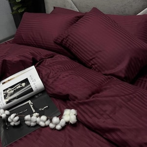 Op de afbeelding: Een bordeauxkleurige beddengoedset met een streepjespatroon. De set bevat kussens en een dekbedovertrek. Een tijdschrift en katoentakjes liggen op het bed. Het beddengoed is gemaakt van een zachte, gladde stof.