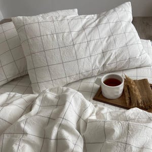 May include: White bedding set with a grid pattern, including pillows and a duvet. A white mug with tea sits on a wooden tray with a decorative element. The bedding has a textured appearance.
