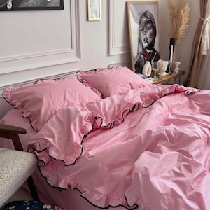 May include: A solid light pink bedding set featuring two pillowcases and a duvet cover, each adorned with ruffled edges trimmed in a thin black border. The fabric has a smooth texture, and the ruffles create a layered, gathered appearance.