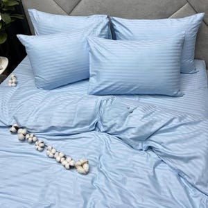 May include: A light blue bedding set featuring a subtle striped pattern, including a duvet cover and four matching rectangular pillowcases. A decorative branch with white cotton bolls is placed diagonally across the lower section of the bed.