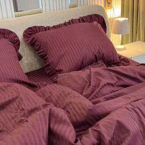 May include: A close-up of a bed with a claret striped bedding set. The set includes a ruffled pillow and a duvet cover. The bedding is made of a soft, silky material. The overall theme is bedroom decor.