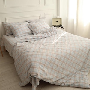 May include: A bed dressed with a light blue, tan, and white plaid bedding set. The set includes a fitted sheet, duvet cover, and two pillowcases. The bed is in a bright room with a white headboard and a record player on a side table.