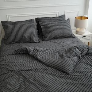 May include: A bedding set featuring a dark gray and black pinstripe pattern. The set includes two rectangular pillows and a matching duvet cover draped over a bed. The fabric has a visible texture, and the stripes run vertically across the pillows.