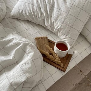 May include: A cozy bedroom scene featuring a white bed with a checkered pattern. A wooden tray holds a cup of tea, dried wheat, and a burlap accent. The bedding includes a pillow and a duvet, creating a relaxing atmosphere.