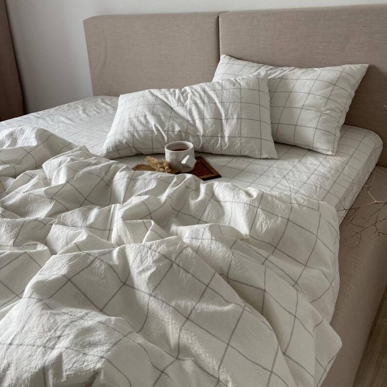 May include: A bed with white bedding featuring a grid pattern. Two pillows and a duvet cover are visible. A small wooden tray with a cup of tea and a snack sits on the bed. The headboard is a neutral beige color.