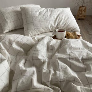 May include: White bedding set with a grid pattern in gray. The set includes two pillows and a duvet cover. A wooden tray with a cup of tea and a snack sits on the bed. The bedding has a soft, wrinkled texture.