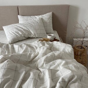 May include: A bed with white bedding featuring a grid pattern. Two pillows rest on the bed, and a cup of coffee sits on a small wooden tray. A woven basket and decorative branches are in the background.