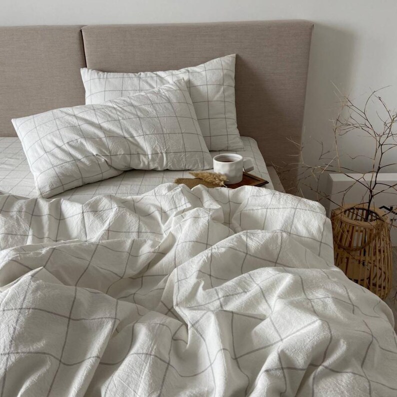 May include: A bed with white bedding featuring a grid pattern. Two pillows rest on the bed, and a cup of coffee sits on a small wooden tray. A woven basket and decorative branches are in the background.