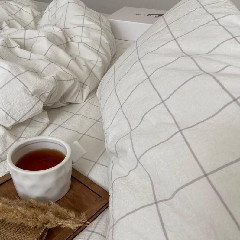 May include: A white ceramic mug filled with tea sits on a small wooden tray, next to dried decorative plants. The mug is on a bed with white bedding featuring a gray grid pattern. A white box is in the background.