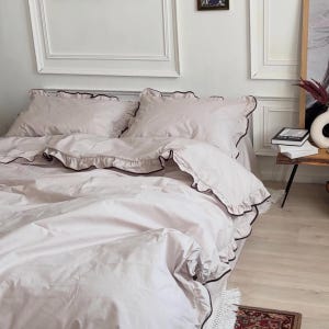 May include: A light beige bedding set featuring a duvet cover and two matching pillowcases. Each piece is adorned with ruffled edges trimmed in a thin, dark brown piping. The fabric appears smooth and solid-colored, creating a uniform appearance.
