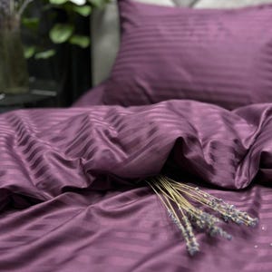May include: A deep purple bedding set featuring a subtle, repeating satin stripe pattern. The set includes a pillowcase and a duvet cover, both displaying the same monochromatic striped texture. A small bundle of dried lavender rests on the fabric.