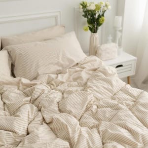 Op de afbeelding: Beige en wit gestreepte beddengoedset, inclusief een dekbed en kussenslopen. De stof heeft een zachte, gekreukte textuur, wat een comfortabele en uitnodigende uitstraling geeft. Het neutrale kleurenpalet en het eenvoudige patroon creëren een rustgevende sfeer.