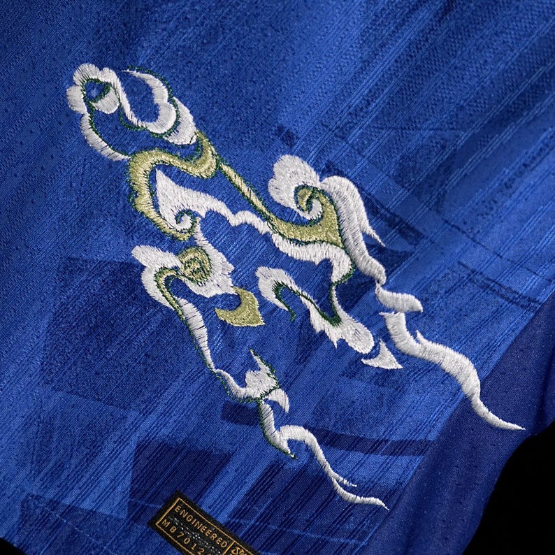 May include: Close-up of a royal blue fabric with an embroidered cloud design in white and gold. The fabric has a textured pattern. The bottom left corner has a tag that reads "ENGINEERED MB7012".