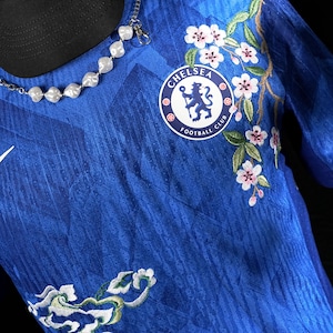 May include: Blue Chelsea Football Club jersey with floral embroidery. The jersey features the team's logo and a white Nike swoosh. A silver necklace with white beads is draped across the jersey.