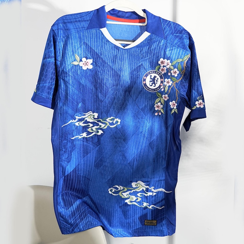 May include: Blue football shirt with white detailing, displaying the Chelsea Football Club emblem and floral embroidery. The shirt has a V-neck collar and short sleeves. Embroidered cloud and floral patterns adorn the front.