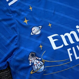 Puede incluir: Camiseta deportiva azul real con rayas y texto blancos. La camiseta presenta parches bordados con tem&aacute;tica espacial, que incluyen planetas, estrellas y un astronauta. El dise&ntilde;o incorpora un sutil patr&oacute;n de rayas.
