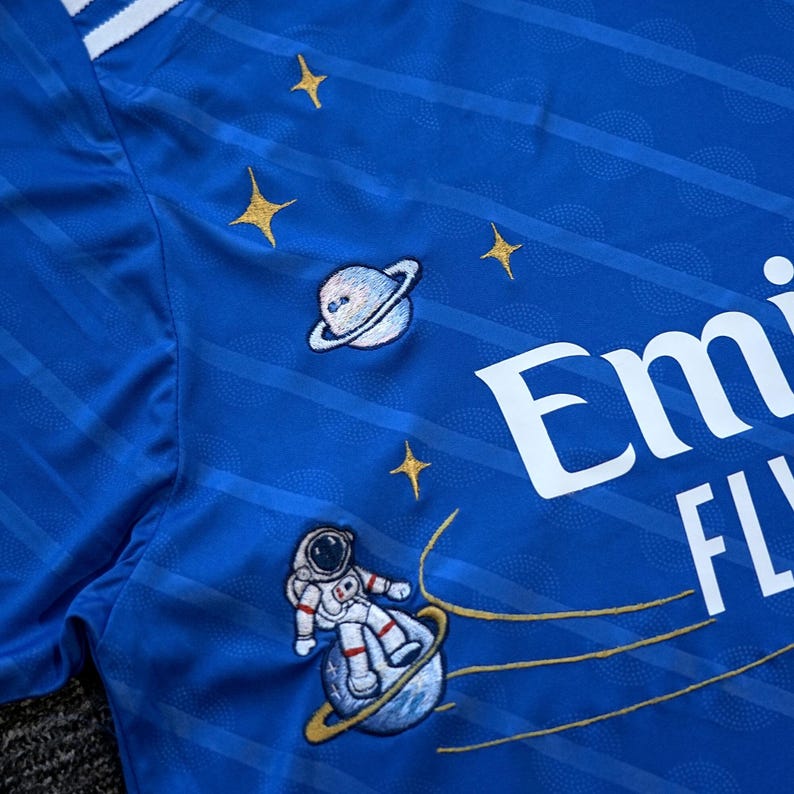 Puede incluir: Camiseta deportiva azul real con rayas y texto blancos. La camiseta presenta parches bordados con tem&aacute;tica espacial, que incluyen planetas, estrellas y un astronauta. El dise&ntilde;o incorpora un sutil patr&oacute;n de rayas.