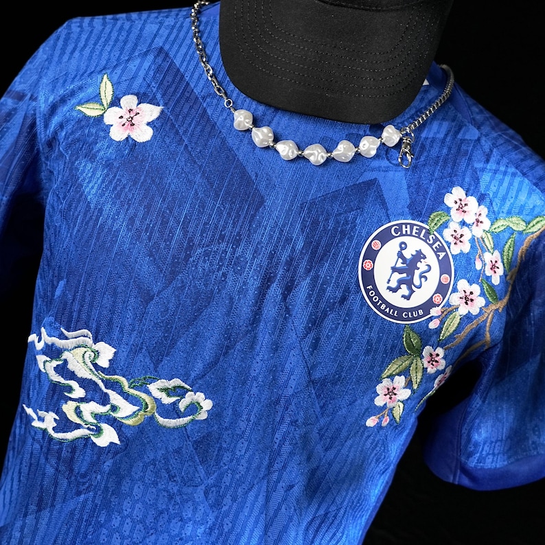 May include: A blue football shirt featuring floral embroidery in white and pink, along with the Chelsea Football Club emblem. A silver chain necklace with white, organic-shaped beads is draped over the shirt. A black baseball cap is partially visible.